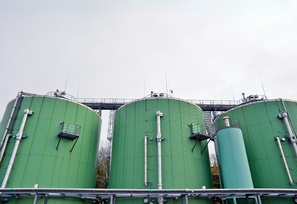 Large green cylindrical anaerobic digestion tanks at a waste treatment facility, featuring industrial pipework, safety railings, and elevated walkways against a grey sky. The tanks are used for processing organic waste into biogas through oxygen-free decomposition.