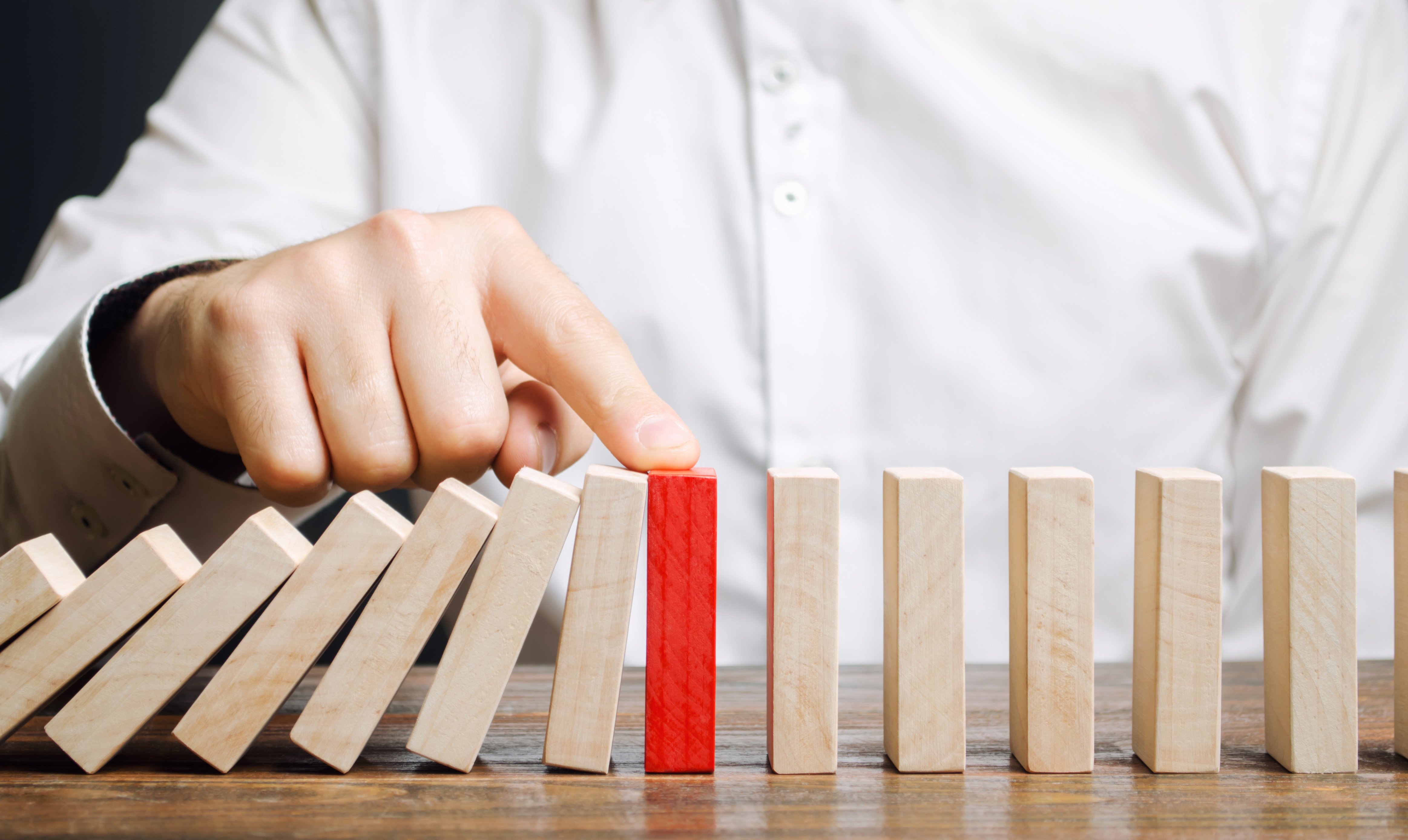 A person in a white shirt stopping a chain reaction of falling wooden dominoes with a red domino piece standing firm in the middle, symbolizing business continuity management and risk prevention. The image illustrates how proper planning can prevent cascading failures in business operations.