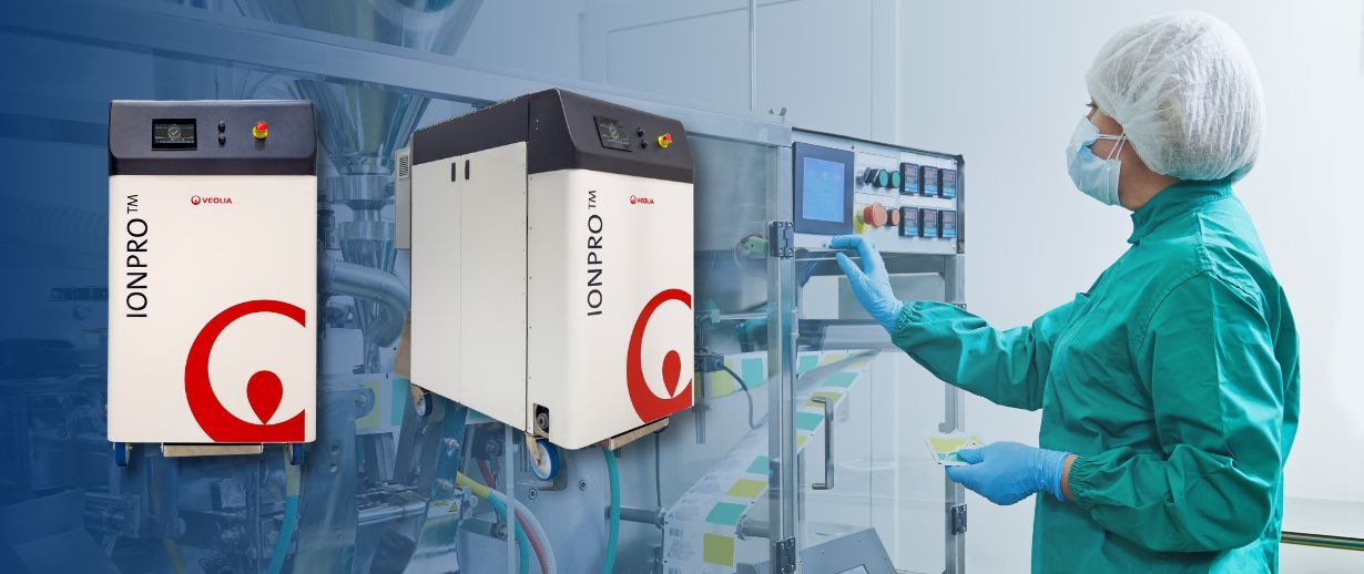 A pharmaceutical technician in protective gear (hairnet, mask, and green gown) operating pharmaceutical technology in a clean room environment. Two white Ionpro units with distinctive red logos are visible, alongside a control panel with digital display.