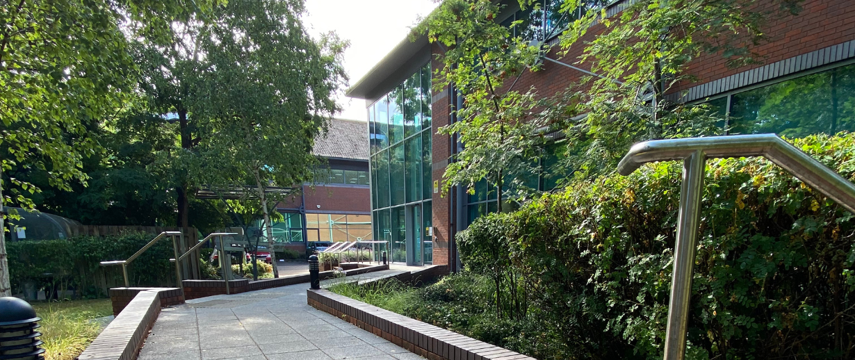 Modern office building surrounded by lush greenery, featuring contemporary glass and brick architecture with accessible ramps and landscaped pathways.