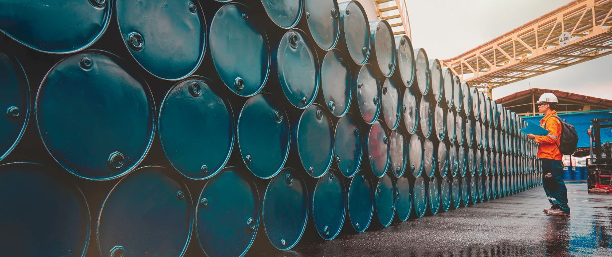 Industrial worker in orange safety vest and hard hat inspecting large stacks of blue chemical storage drums at a treatment facility. The drums are neatly arranged in rows on a wet concrete surface under an industrial structure